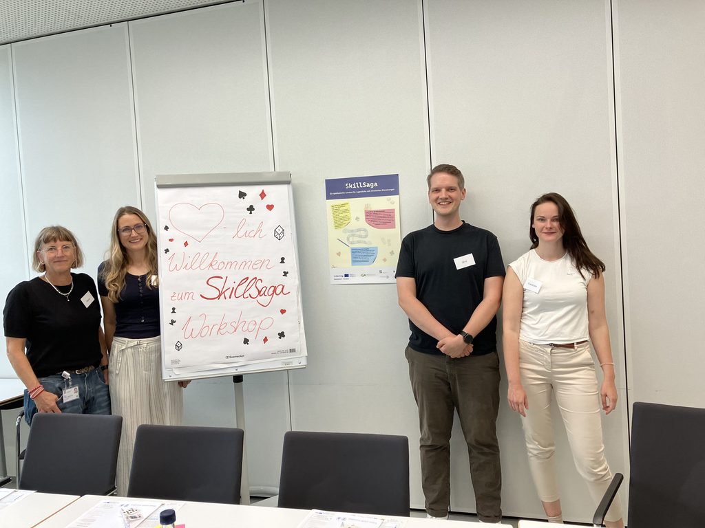 Four people pose beside a flip chart reading “Herzlich willkommen zum SkillSaga Workshop.”