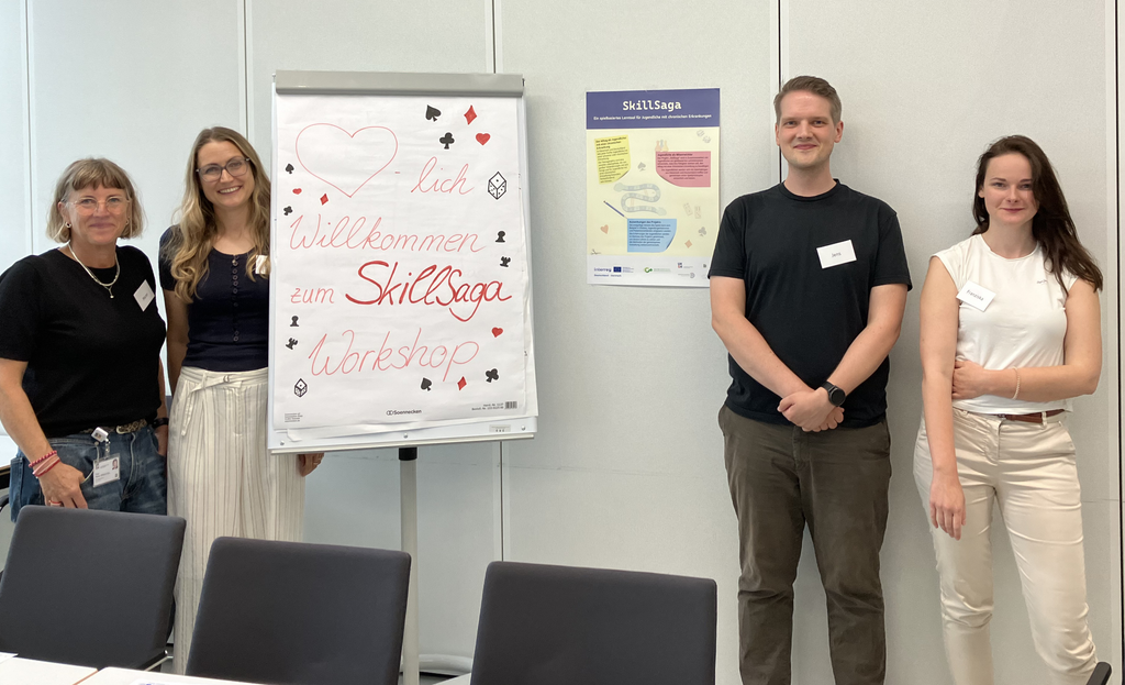 Four members of the SkillSaga team standing next to a flip chart with a hand-drawn welcome to the SkillSaga workshop and a SkillSaga poster on the wall