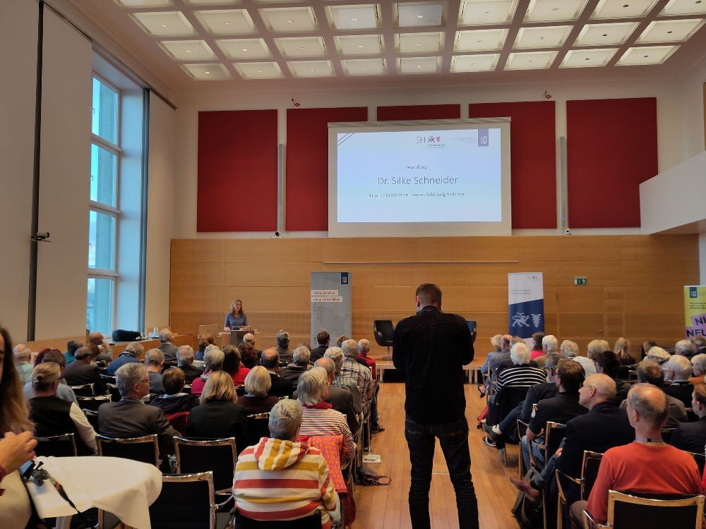 Dr. Silke Schneider speaking at a lectern in a full hall in the Schleswig-Holstein State Parliament, her name shown on the projection screen behind her